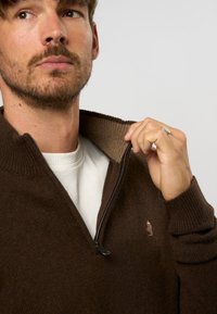 Rudas zip-up megztinis su raukšlėtu apykakle ir rankogaliais, turintis kontrastingą šviesiai rudą vidinę apykaklę ir mažą logotipą ant krūtinės.