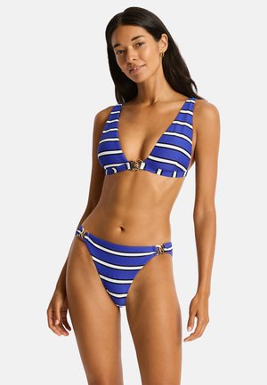 Kobieta w niebiesko-białym paskowanym bikini ze złotymi klamrami na białym tle.