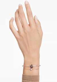 Bracelet manchette en or rose avec un design de cygne, accents noirs et une petite pierre claire, porté sur une main à la peau claire.