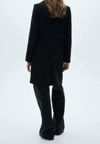 Manteau noir sur mesure avec une texture lisse, présentant une coupe droite et des manches longues, associé à un pantalon noir ample.