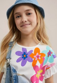 Witte t-shirt met grote, kleurrijke bloemenprints in paars, oranje, roze en blauw, gecombineerd met denim overall en een lichtblauwe emmerhoed.