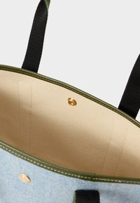 Detailní záběr na otevřenou světle modrou tašku přes rameno s olivově zeleným lemováním, béžovou látkovou interioru, černými uchy a zlatým patentním knoflíkem.