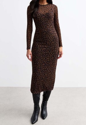 Nainen, jolla on päällään vartalonmyötäinen, pitkähihainen, nilkkaan ulottuva leopardi-kuvioinen mekko ja mustat polvenkorkuiset teräväkärkiset saappaat, seisoo harmaalla lattialla.