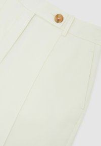 Pantalon couleur crème en tissu lisse, doté d'un grand bouton marron unique, de plis et de poches latérales.