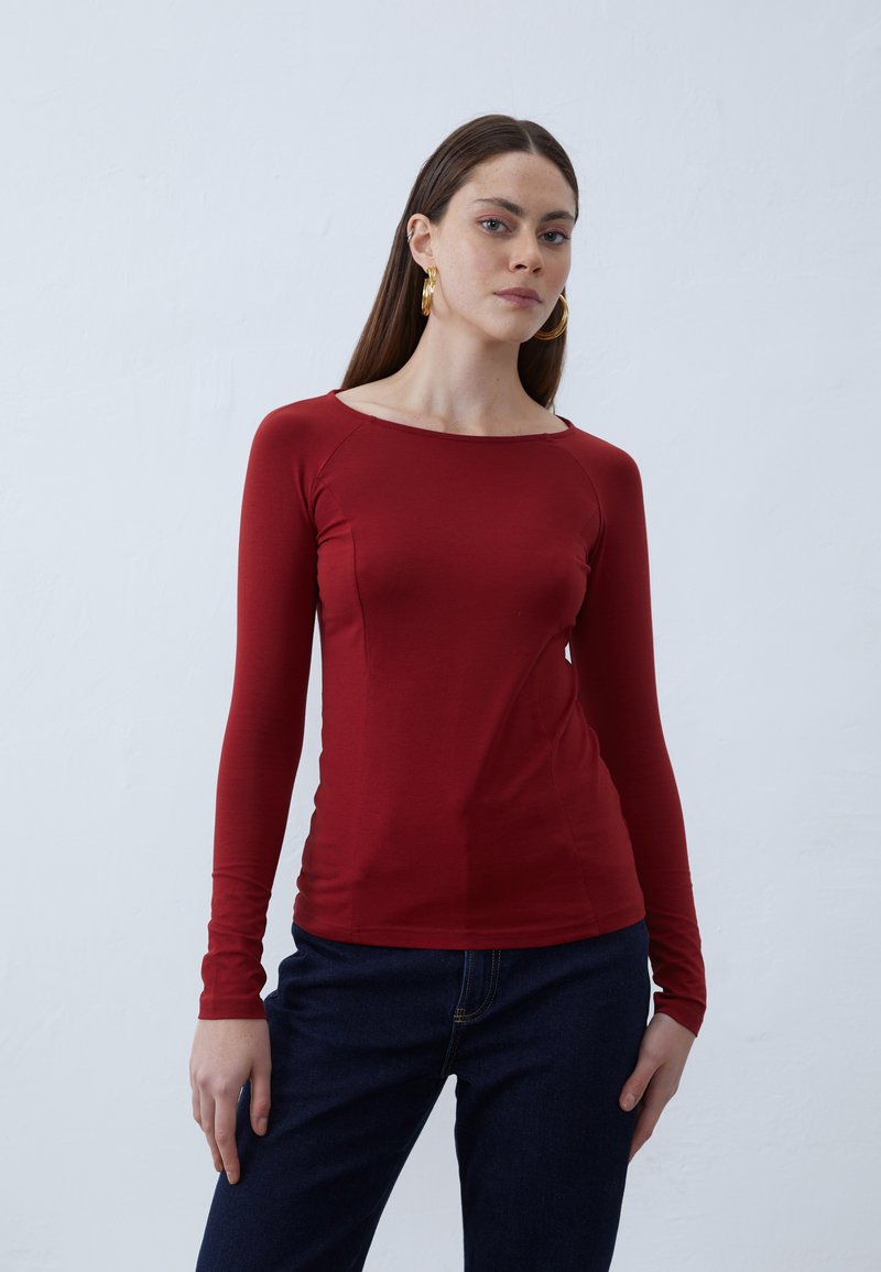 Red long-sleeve top with a bateau neckline, made from a smooth, stretchy fabric. Paired with dark blue jeans, displayed against a white background.