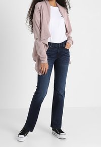 Hellrosa offener Cardigan mit drapiertem Vorderteil, getragen über einem weißen T-Shirt. Dunkelblaue Jeans und schwarze Turnschuhe vervollständigen das Outfit.