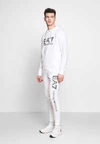Λευκή μπλούζα και αντίστοιχες φόρμες σε λευκό χρώμα με μαύρο κείμενο "EA7 Emporio Armani". Λείο ύφασμα, εφαρμοστό σχέδιο και ελαστικές μανσέτες. Λευκά αθλητικά παπούτσια.
