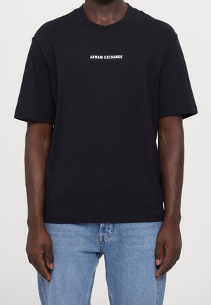 Mężczyzna w czarnej koszulce z krótkim rękawem z białym napisem "ARMANI EXCHANGE" i jasnoniebieskich dżinsach na jednolitym tle.