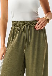 Pantaloni a gamba larga color verde oliva in tessuto strutturato, con vita elasticizzata e coulisse regolabile. Mano visibile che indossa un braccialetto.