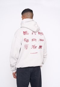 Svetlošedý oversize sweatshirt s viacerými červenými logami a textom na chrbte, rebrovanými manžetami a veľkou kapucňou. Tkanina z bavlnenej zmesi.