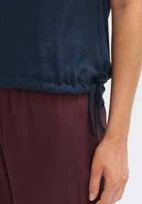 Top a costine blu navy con un cordoncino all'orlo, abbinato a pantaloni bordeaux. Il tessuto appare morbido e con texture.