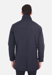 Giacca isolante blu navy con colletto alto, taglio dritto e maniche lunghe. Presenta una texture liscia e un dettaglio sullo spacco posteriore.