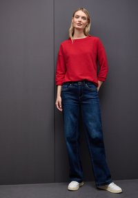 Top rosso a maniche lunghe e costine abbinato a jeans ampi blu scuro. L'outfit è completato da sneakers bianche. Sfondo neutro.