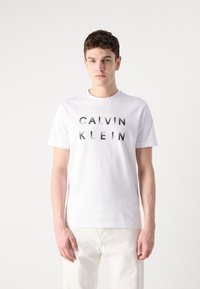 White cotton T-shirt featuring black printed text "CALVIN KLEIN" in a bold, modern font. Crew neckline and short sleeves.