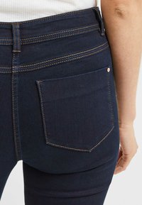 Jeans in denim blu scuro con vita alta, cuciture arancioni e una sola tasca posteriore con un piccolo dettaglio del marchio. Texture liscia.