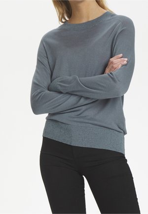 Mujer con suéter azul de manga larga, brazos cruzados y pantalones negros, sobre un fondo blanco liso.
