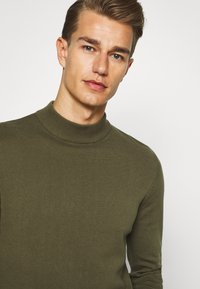 Suéter de cuello alto verde oliva hecho de un tejido de punto texturizado con un diseño ajustado, que presenta un cuello de canalé y detalles de hombros sin costuras.