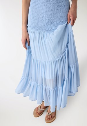 Donna che indossa una gonna lunga a balze azzurro chiaro con vita arricciata, solleva leggermente l'orlo per mostrare sandali beige con cinturini metallici.