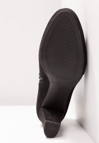 Bottine femme en daim noir appuyée contre un mur, montrant la semelle texturée et le talon bloc avec le texte « SMALL SHOE » sur l'extrémité du talon.