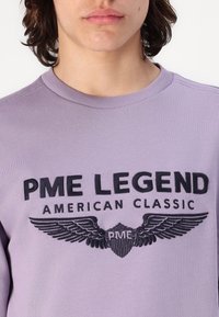 Sudadera de color lila claro con cuello redondo acanalado, que presenta un destacado estampado negro con la frase "PME LEGEND" y un diseño gráfico de alas. Textura suave.