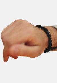 Schwarzes Perlenarmband aus mattem Stein mit metallischen Akzenten, getragen an einer Hand, die eine Faust bildet. Es verfügt über unterschiedliche Perlengrößen und -strukturen.