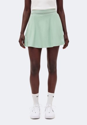 Kobieta ubrana w jasnografitową sportową spódnicę, biały bezrękawnik, białe skarpetki Nike i białe tenisówki na sznurowadła, stojąca na jednolitym tle.