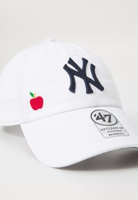 Gorra de béisbol blanca con logotipo de NY en azul marino, emblema de manzana roja, correa ajustable y etiqueta con la marca "47 Clean Up". Material de algodón.