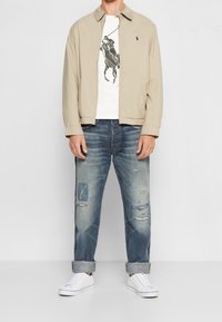 Béžová lehká bunda přes bílé tričko s barevným logem koně, doplněná o potrhané modré džíny a bílé tenisky.