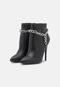 Botas de cuero negro hasta el tobillo con puntera afilada y tacones de aguja, con un detalle de cadena plateada alrededor del tobillo. Textura suave y diseño elegante.