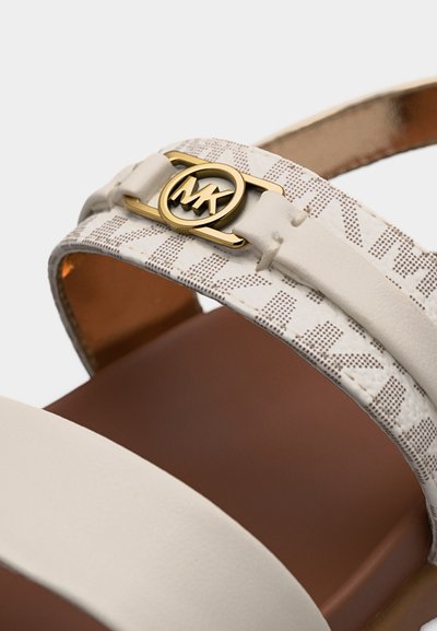 Λευκό λουρί σανδάλου με μπεζ διακοσμητικό περίβλημα, χρυσή κυκλική αγκράφα με το λογότυπο MK και καφέ σόλα ορατή στο φόντο.