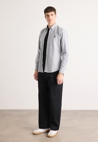 Lichtgrijze button-up shirt met een puntige kraag, zwarte gelaagde t-shirt eronder, gecombineerd met een relaxte zwarte broek en witte sneakers.