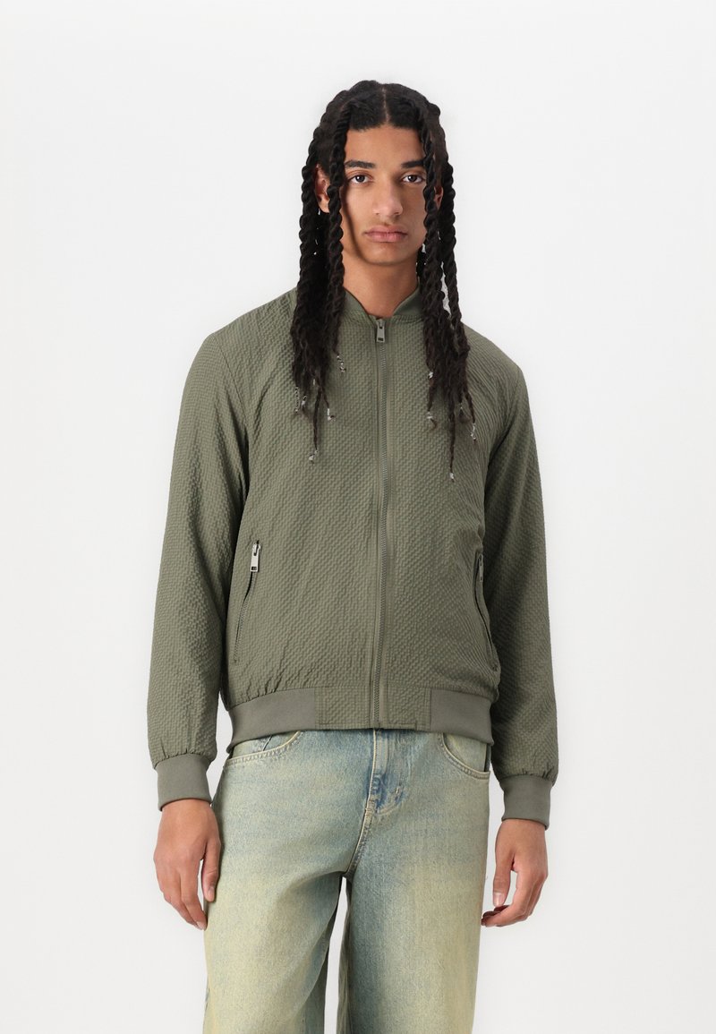 Olive green bomber jacket with textured fabric, fitted cuffs, and zip-up front. Features side pockets and a simple collar design.