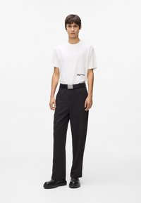 Jauns vīrietis stāv saskatē, ģērbies baltā Karl Lagerfeld T-kreklā, melnās biksēs ar jostu un melnās kurpēs uz vienkārša baltā fona.