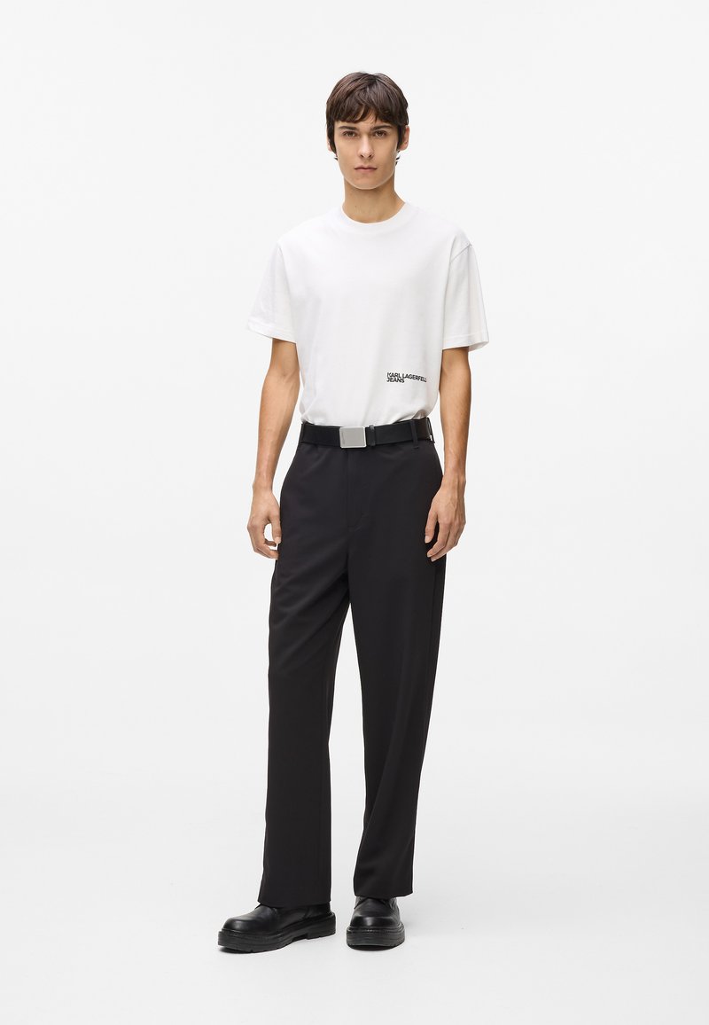 Jauns vīrietis stāv saskatē, ģērbies baltā Karl Lagerfeld T-kreklā, melnās biksēs ar jostu un melnās kurpēs uz vienkārša baltā fona.