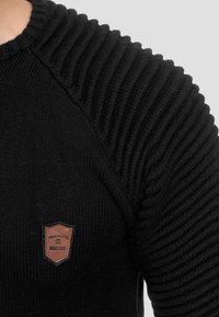 Pull noir en tricot avec une texture côtelée sur les épaules, arborant un écusson marron sur la poitrine portant l'inscription "WEAR THE CODE."