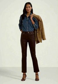 Ženska v temno modri denim srajci, rjavih hlačah, črnih petah, ki drži bež suknjič čez ramo in stoji ob enobarvni steni.