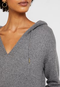 Grijze gebreide hoodie met een V-hals, trekkoorddetails en geribbelde accenten. Heeft een zachte textuur en een ontspannen pasvorm.