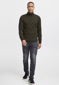 Pulover cu guler înalt de culoare verde închis, cu manșete și tiv cu model canelat, asortat cu blugi negri decolorati și adidași negri cu accente albe.