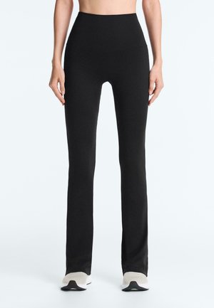 Žena oblečená v čiernych legínach s vysokým pásom a rovnými nohavicami a bielych teniskách, stojaca na jednoduchom svetlom pozadí.