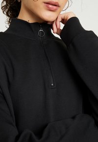 Felpa nera con zip, colletto alto, polsini a coste e una cerniera decorativa. Texture liscia e vestibilità ampia, visibile dalla vita in su.