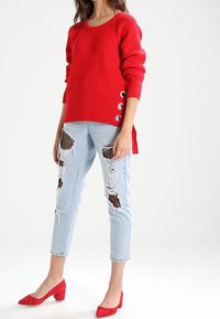 Red ribbed sweater with side button accents, paired with light blue distressed jeans featuring mesh patches, and red high-heeled shoes.