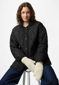 Giacca nera trapuntata con bottoni a pressione, accompagnata da guanti a coste color crema. Il modello indossa un maglione beige sotto. Visibili jeans blu scuro.