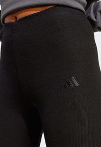 Črne rebraste legice z visokim pasom, na spodnji levi strani z vezenim črnim logotipom Adidas. Mehka tekstura, tesno prilegan dizajn.