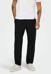 Sorte bukser med en afslappet pasform, der har to forlommer, lavet af glat stof, kombineret med hvide sneakers og en simpel hvid t-shirt.