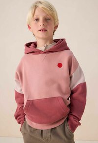 Sweat-shirt à capuche en nuances de rose avec un design en coloris bloqués, style pull, poche avant et un petit logo en forme de cercle rouge.