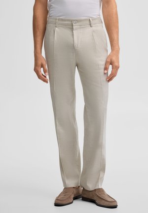 Mann trägt beige maßgeschneiderte Hose, weißes eingestecktes Hemd und braune Wildleder-Loafer, steht vor einem schlichten hellen Hintergrund.