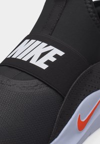 Κοντινό πλάνο μαύρου αθλητικού παπουτσιού Nike χωρίς κορδόνια με λευκό λογότυπο Nike σε ελαστικό λουράκι και πορτοκαλί σημαία στο λευκό πέλμα.