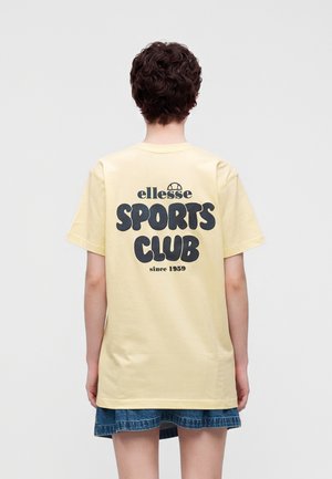 Personne aux cheveux courts et bouclés, portant un t-shirt jaune pâle avec l'inscription « ellesse SPORTS CLUB since 1959 » et une jupe en jean courte, de dos.