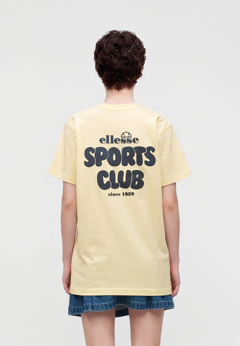 Personne aux cheveux courts et bouclés, portant un t-shirt jaune pâle avec l'inscription « ellesse SPORTS CLUB since 1959 » et une jupe en jean courte, de dos.