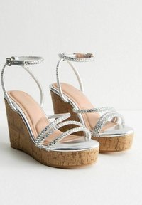 Sandalias de cuña plateadas con múltiples diseños de tiras, plataforma de corcho y acentos metálicos brillantes. Textura suave y estilo de punta abierta.